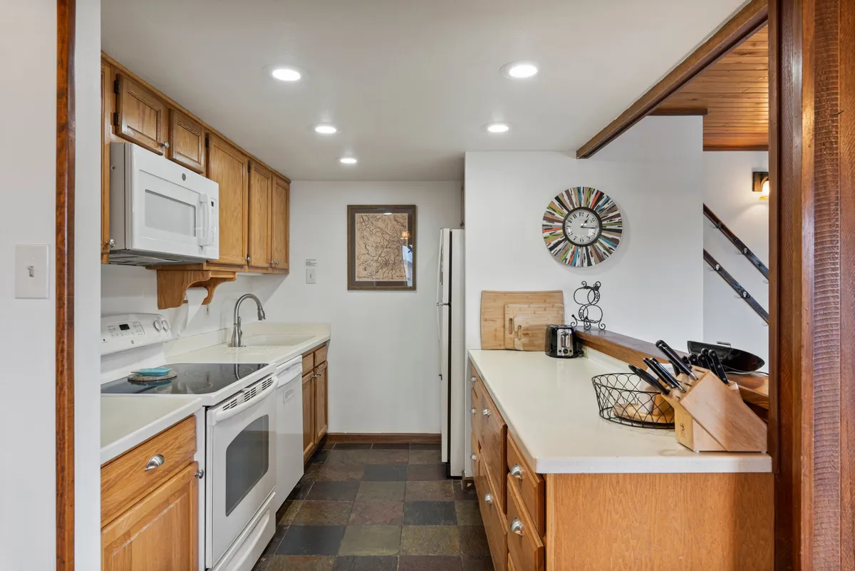 A warmly lit, fully equipped kitchen featuring a spacious countertop, stainless steel refrigerator, clock on the wall, and framed artwork. The countertop is clear and inviting, suggesting plenty of room for meal preparation and social cooking. The overall mood is welcoming, ideal for family gatherings or hosting friends.