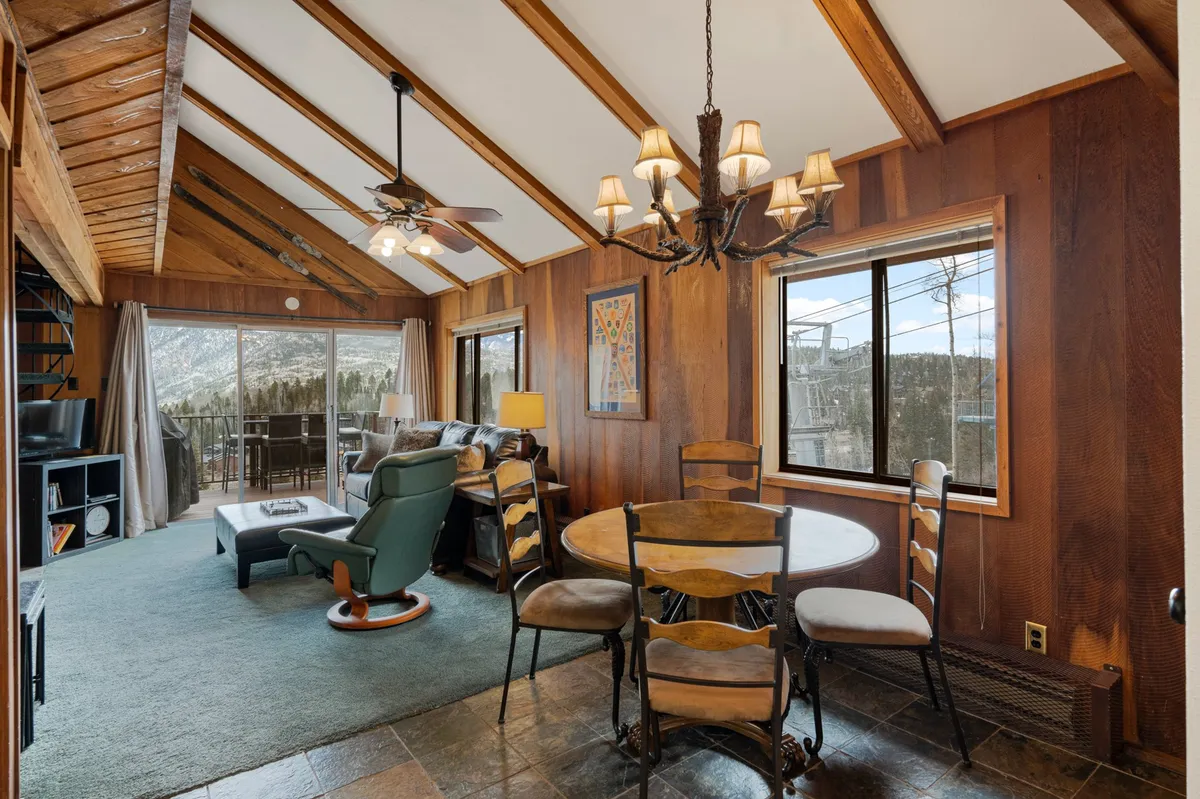 A warm, inviting dining area with a wooden dining table, elegant chandelier, recliner, and windows that offer views of the landscape. The table is set for a meal, encouraging shared moments, game nights, and lingering conversations beneath the soft glow of evening light.