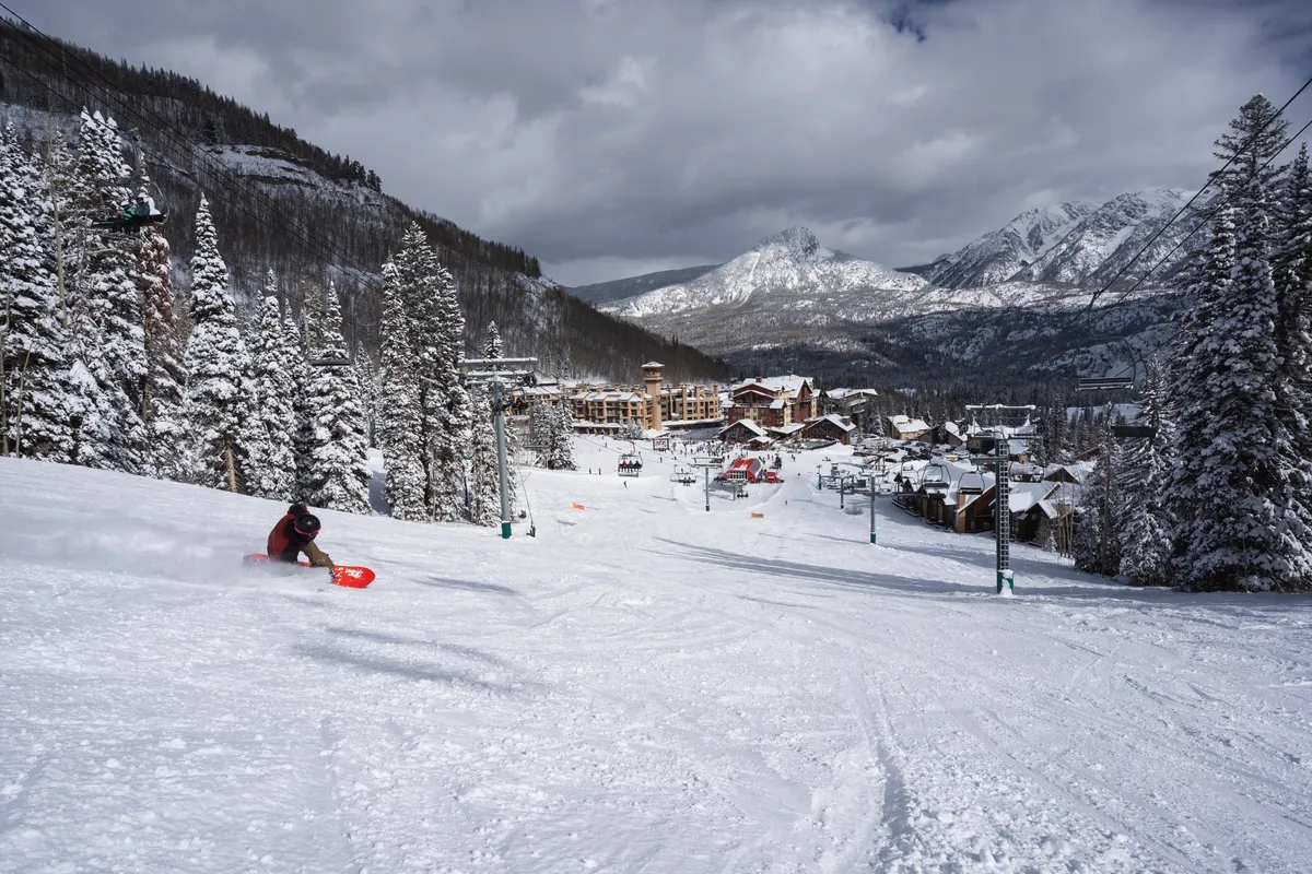 A dynamic scene featuring a skier carving down a pristine, snow-covered slope with the resort buildings and majestic mountain backdrop. The lively winter setting highlights both action and the natural beauty of the location.