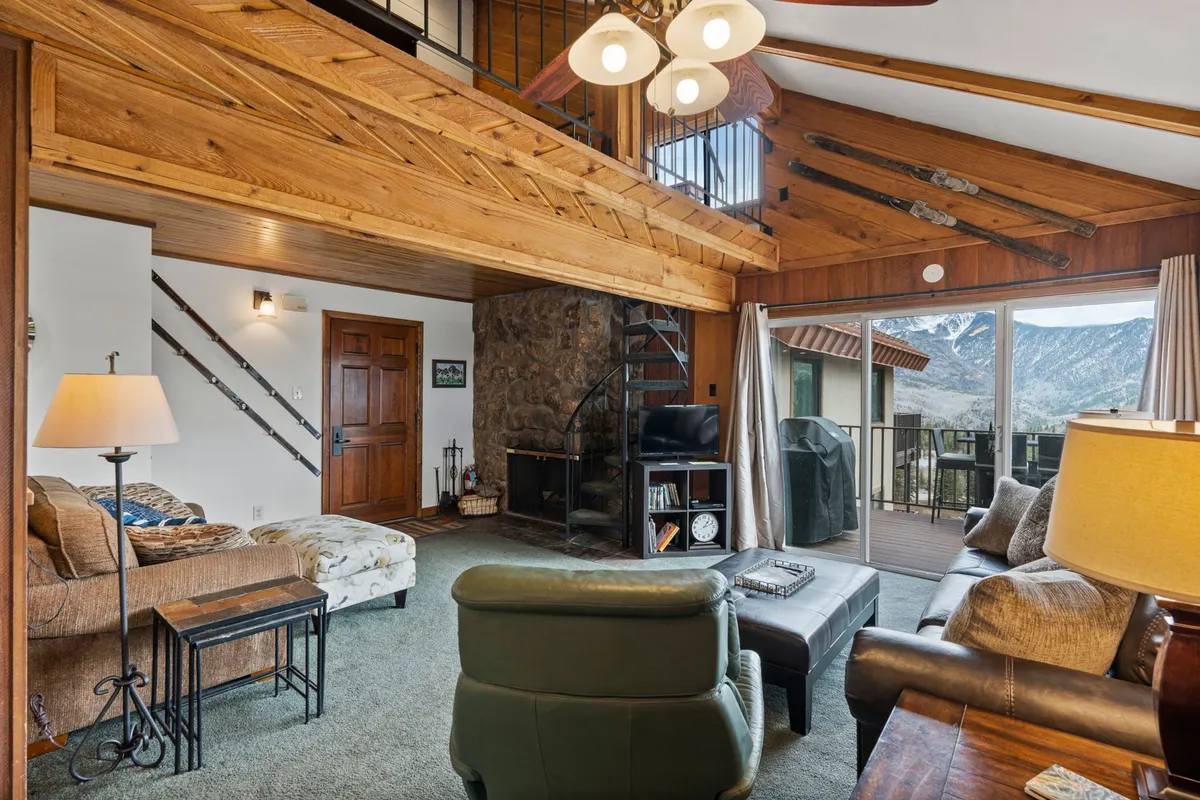 A spacious living room featuring exposed wooden beams, a striking stone fireplace, a spiral staircase, and a comfortable seating area. Large windows frame panoramic mountain views, creating a seamless connection to the outdoors. The scene invites relaxation by the fire or gathering for conversation with friends and family.