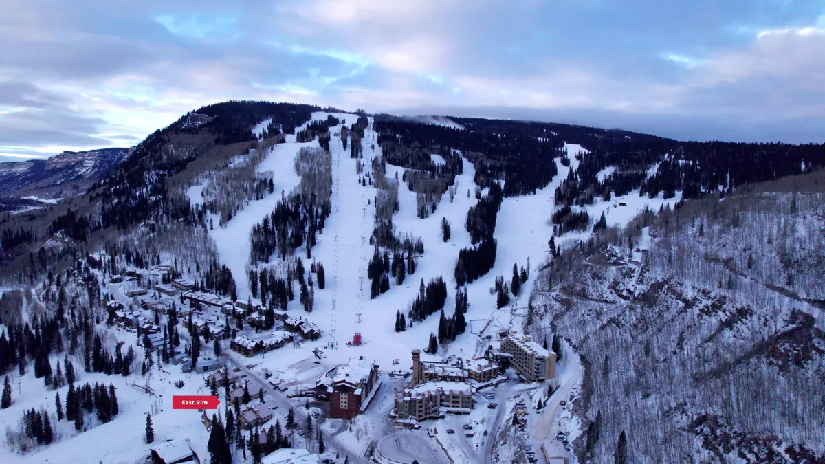 A sweeping view of the resort buildings nestled at the foot of snow-covered ski slopes, with skiers and snowboarders in action and tall trees dusted in fresh powder. The early morning light creates a cozy, adventurous atmosphere, capturing both the excitement of the slopes and the inviting ambiance of the resort.