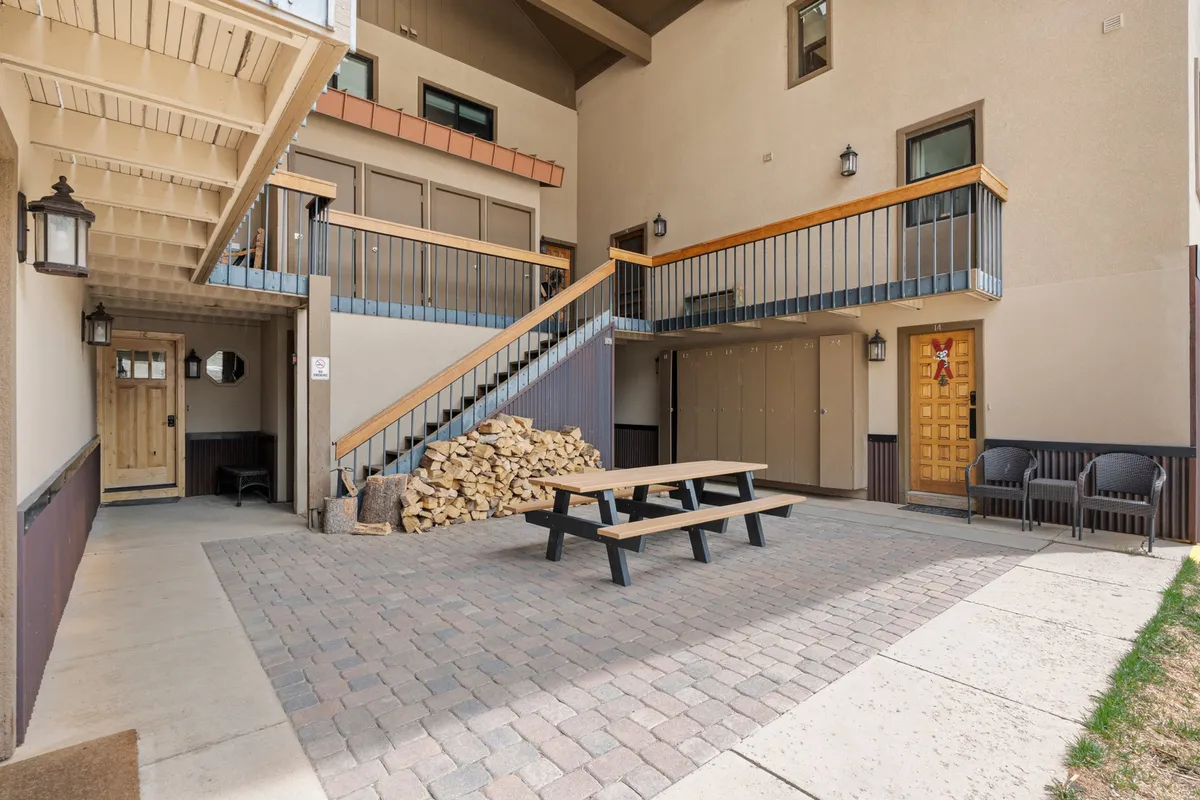 The image features an inviting exterior patio with a wooden picnic table, a stack of firewood, and the welcoming front entrance. The setup suggests enjoying meals outdoors, intimate conversations, and cozy evenings after a day of exploration.