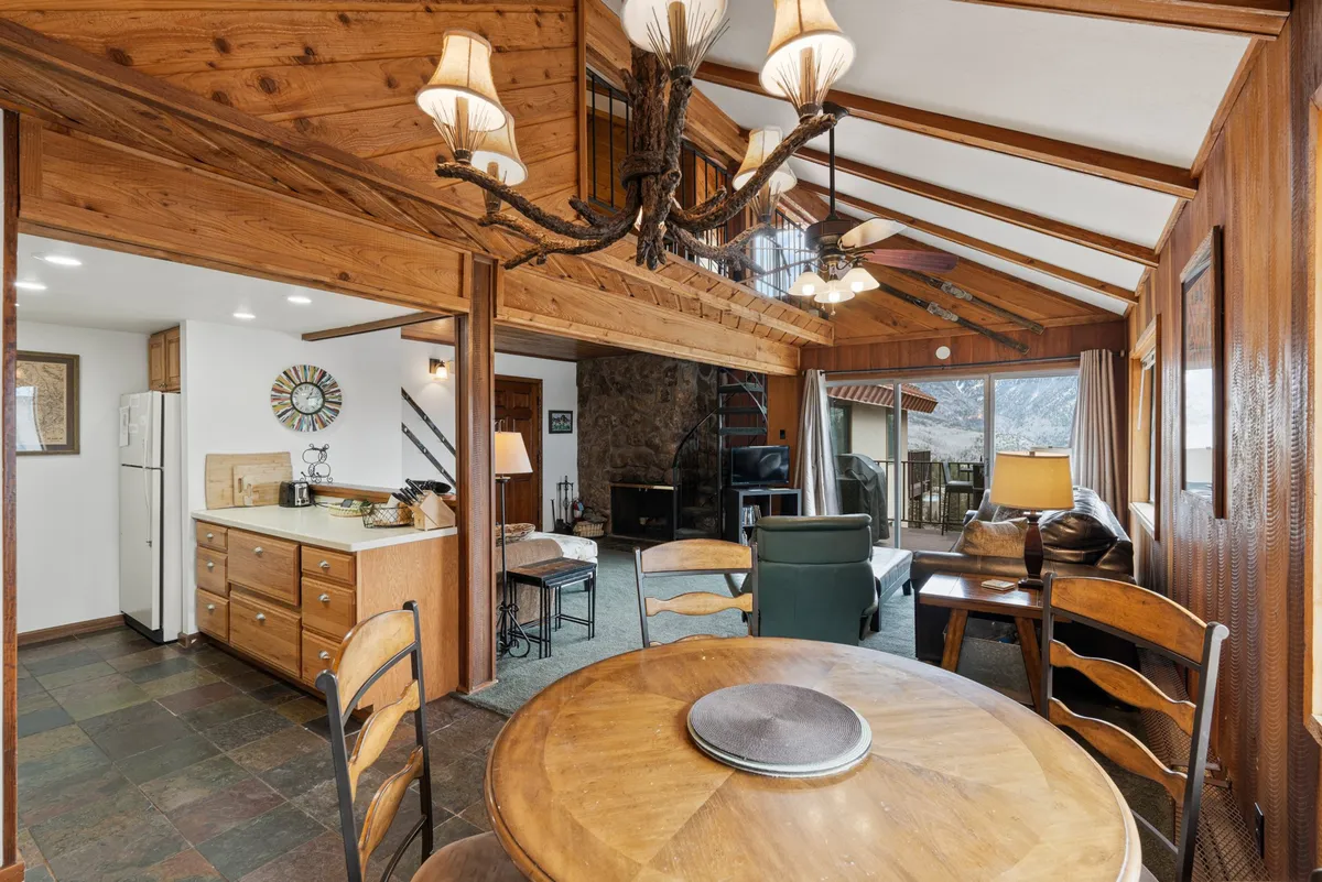An open-plan dining and living area with a rustic wooden chandelier above a dining table, a stone fireplace, and sliding glass doors leading onto a balcony. Ample natural light fills the room, suggesting mountain views and a seamless indoor-outdoor connection.