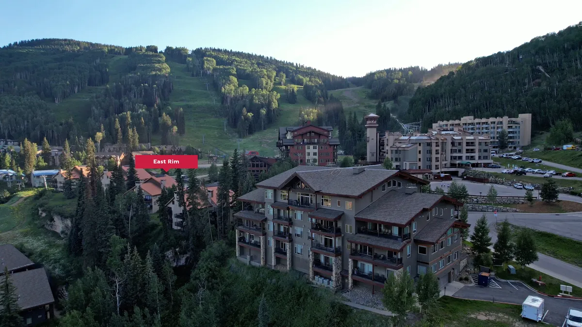This exterior image captures a breathtaking view of the mountains behind residential buildings, with a tree line in the foreground and a parking area that hints at easy resort access. The scenery evokes both serenity and adventure, perfectly setting the mood for a mountain retreat.