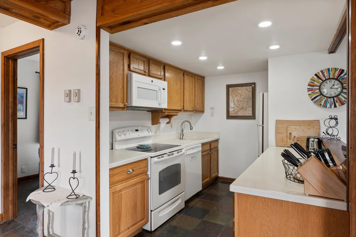 A cozy and functional kitchen scene with warm lighting, wooden cabinetry, a clean countertop, sink, various kitchen utensils, and a wall clock. The setup emphasizes the everyday practicality and functionality of the space, suggesting it's ready for meal prep and casual entertaining.