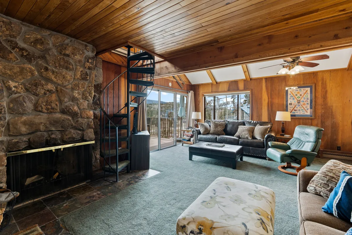 A cozy living room featuring a stone fireplace, a striking spiral staircase, large windows framing mountain views, and an inviting seating area. The space exudes warmth, with natural wood accents and comfortable furniture perfect for relaxation after a day on the slopes.