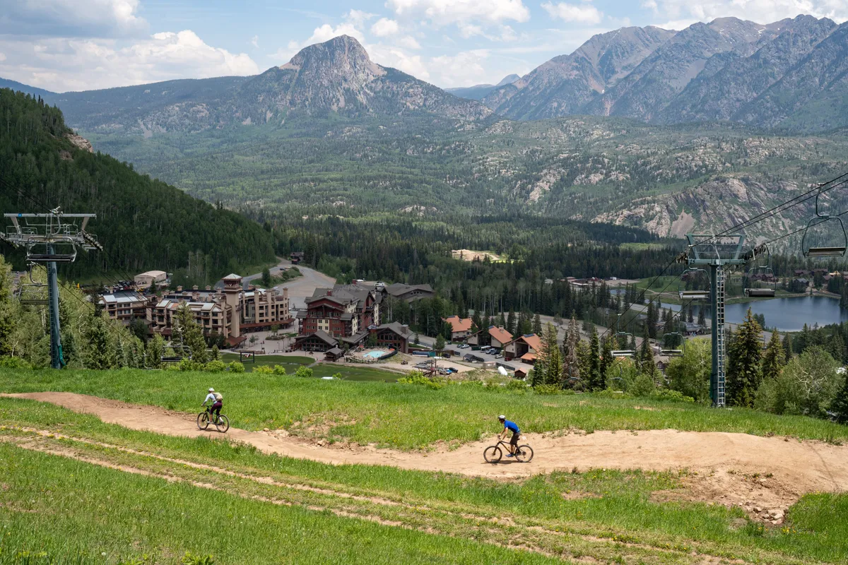 A vibrant exterior view featuring a sweeping mountain range, bike riders traversing trails, resort buildings nestled at the base, a ski lift, and a sparkling lake. The landscape conveys four-season excitement with options for mountain biking, hiking, skiing, and lakeside relaxation.