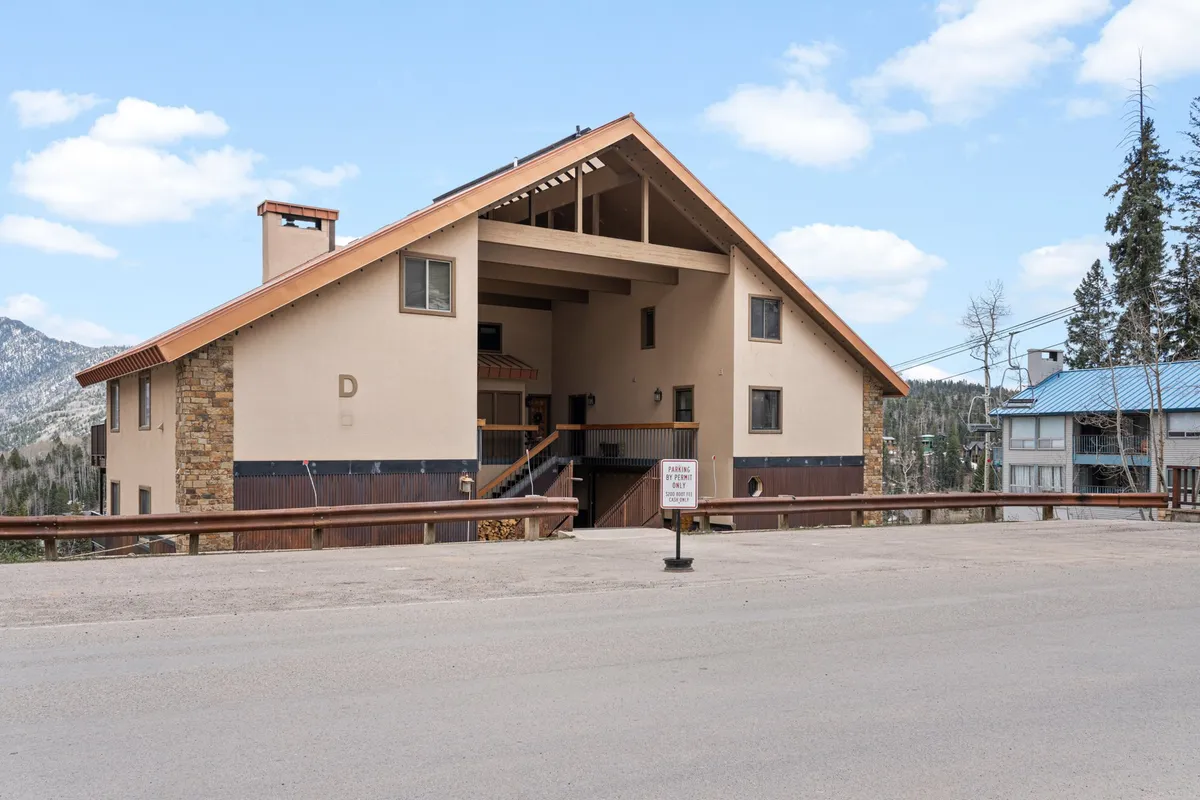 The image showcases the inviting exterior of the East Rim D24 condo in Durango, Colorado, emphasizing its distinctive A-frame architecture, spacious front porch with stone detailing, and the warm glow of sunset casting a cozy, welcoming atmosphere. The porch invites guests to imagine relaxing after a day on the slopes, gathering with family or friends, and soaking in the scenic mountain setting.
