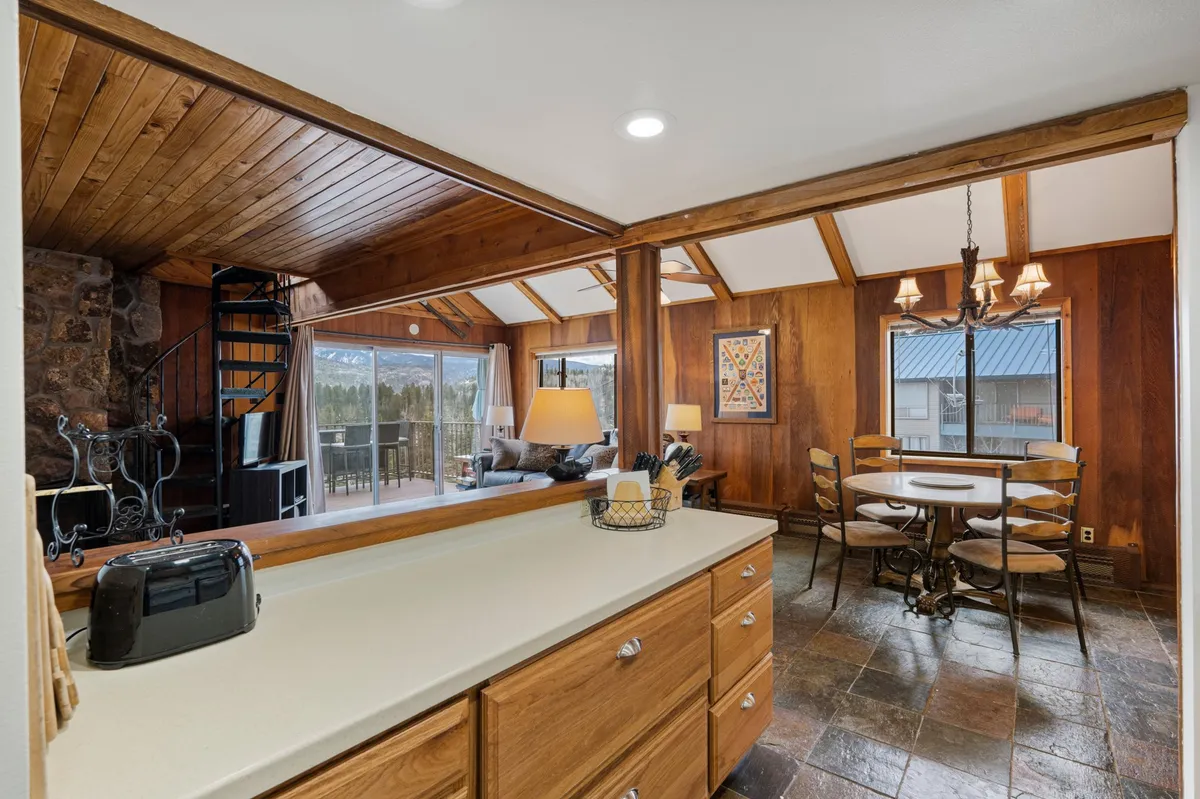 A perspective that includes the kitchen countertop, a spiral staircase, a dining table with chairs, and large windows with mountain views. This open-concept layout visually connects the kitchen and dining area, inviting shared meals and conversations. Natural light and scenic views enhance the welcoming atmosphere.