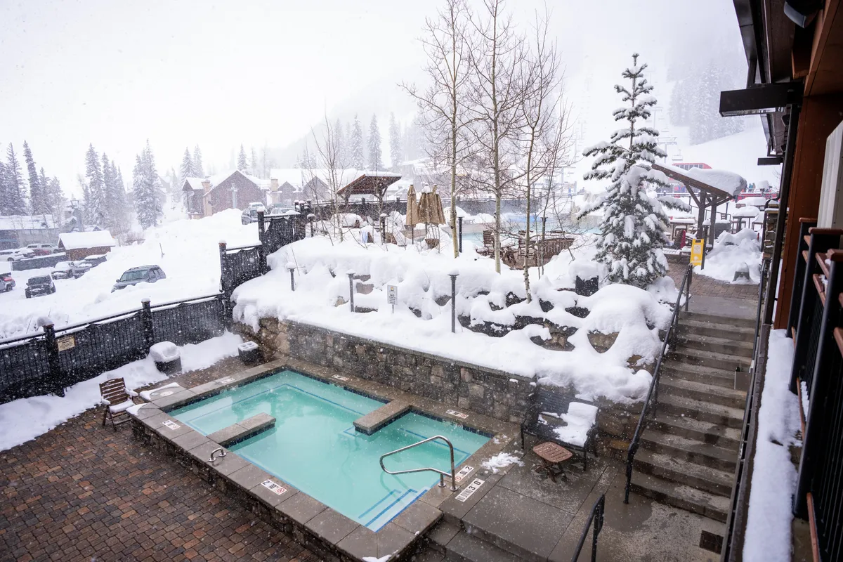 A snow-covered exterior with an outdoor hot tub surrounded by trees and building structures, inviting relaxation in a tranquil, wintry setting. The contrast between the warm water and snowy landscape hints at unique, memorable moments for couples.